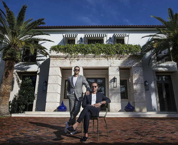 Two luxury real estate brothers posing confidently outside a large upscale villa amid a sunny setting.
