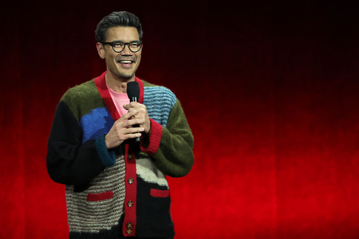 Man wearing glasses and a multicolored sweater speaking on stage, with fans debating Spider-Man Brand New Day changes.