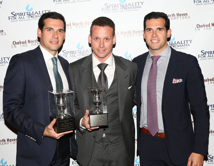 Three men in suits holding trophies at a luxury event, linked to infamous luxury real estate brothers case.