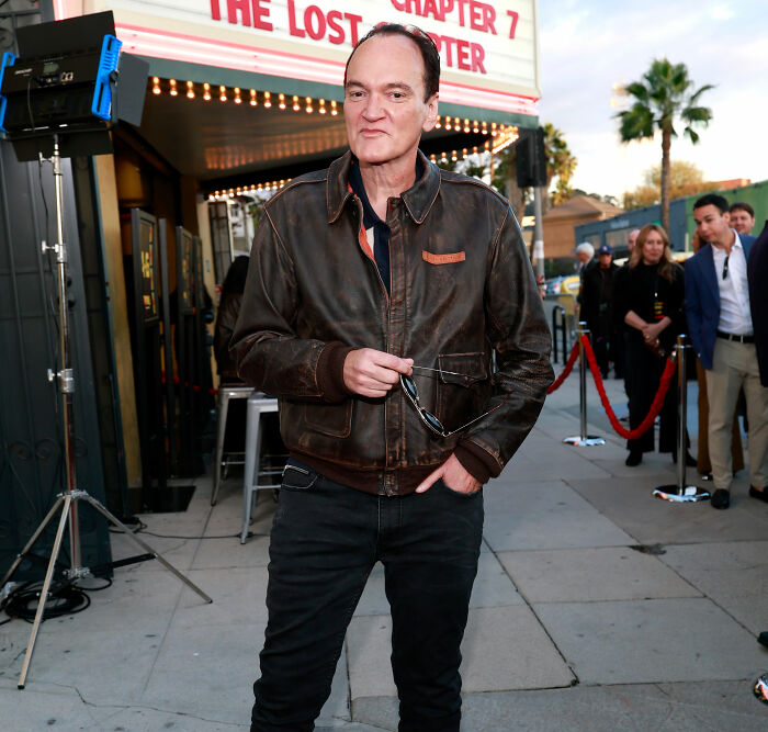 Quentin Tarantino in a brown leather jacket holding sunglasses outside theater amid fans reacting to Rosanna Arquette.