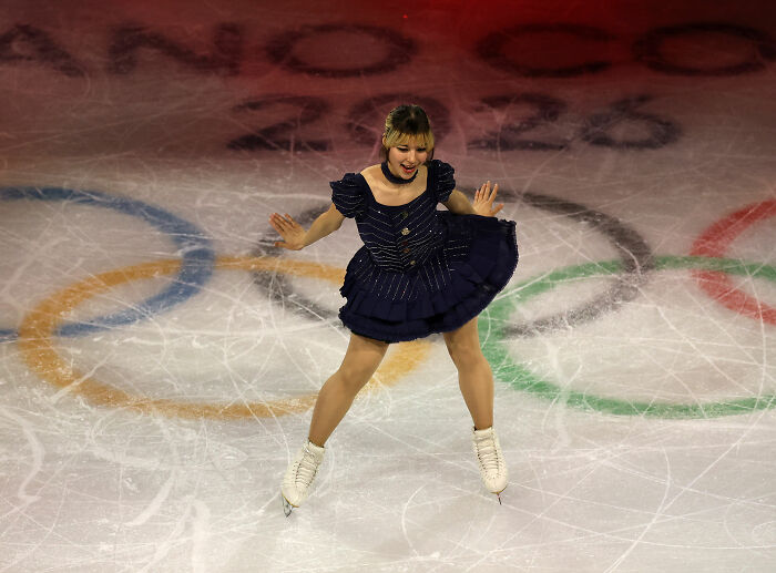 Alysa Liu figure skating gracefully on ice with Olympic rings in background during international competition.