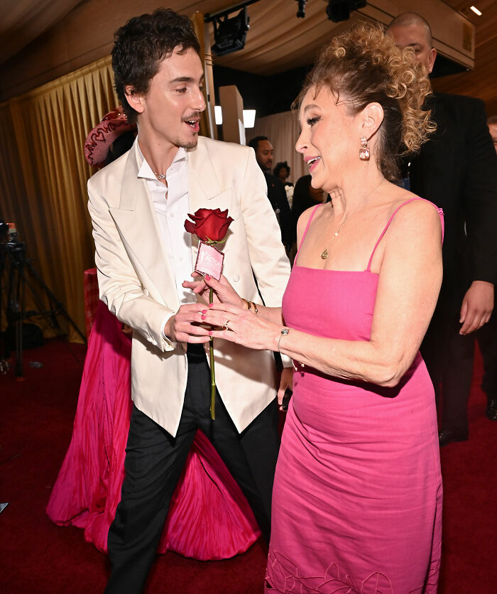 Timoth&eacute;e Chalamet dressed in white giving a rose to a woman in a pink dress at the Actor Awards event.