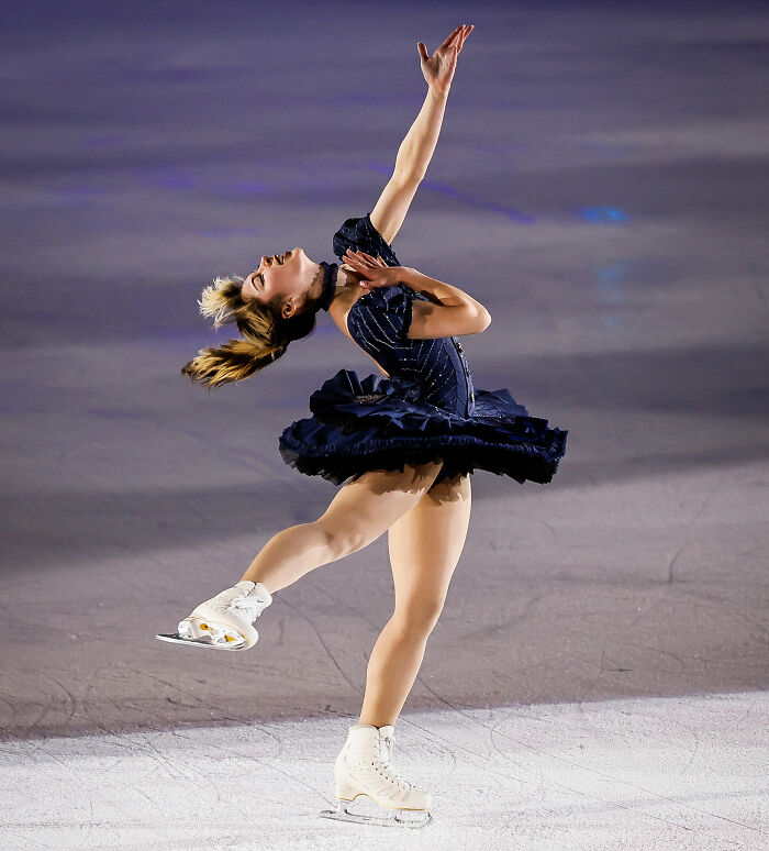 Alysa Liu performing a graceful spin on ice wearing a dark blue dress during a figure skating routine.