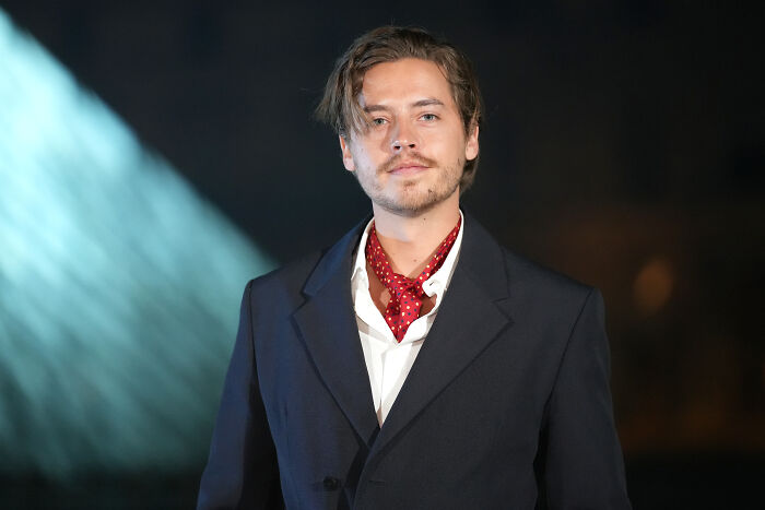 Actor Dylan Sprouse wearing a dark blazer and red scarf at an event, linked to Miley Cyrus breakup news.
