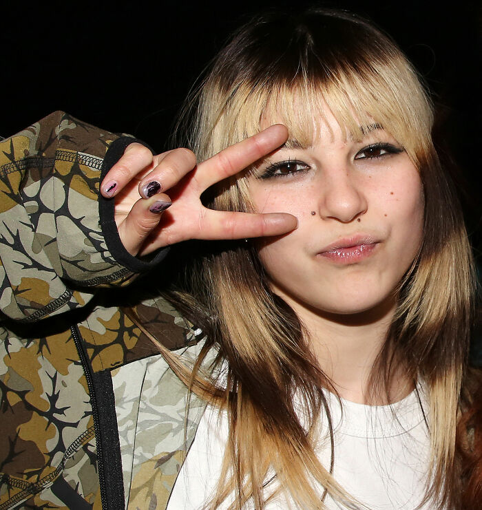 Young woman with blonde and brown hair making a peace sign, representing Olympic hero Alysa Liu and figure skating challenges.
