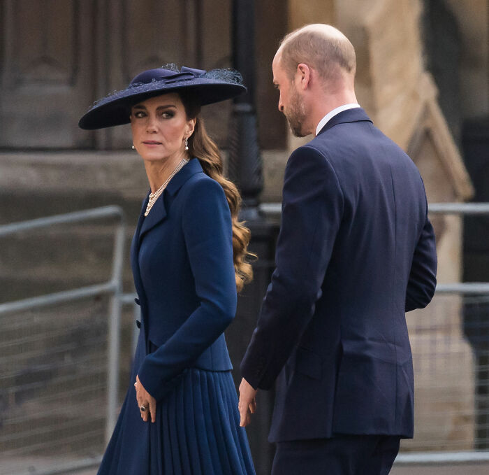 Prince William and Kate Middleton dressed in navy blue attending a royal family gathering with tense expressions.