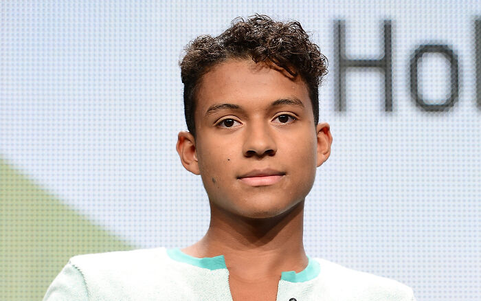 Young male actor from the Michael movie cast wearing a light shirt, posing against a bright, blurred background.