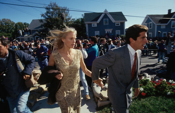 Daryl Hannah in a patterned dress walking with a man in a suit among a crowd, highlighting her JFK Jr. connection.