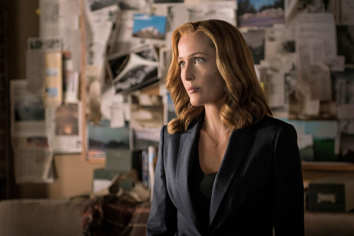 Woman with red hair in a dark blazer stands in front of a wall covered with photos and papers in The X-Files reboot scene.