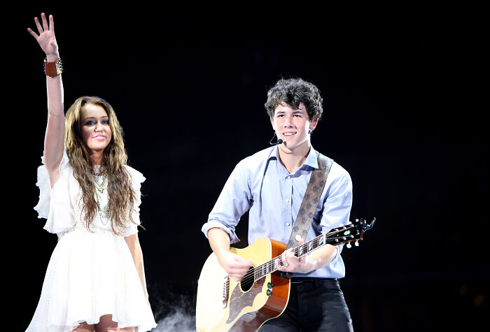 Miley Cyrus waving in white dress beside Dylan Sprouse playing guitar on stage during live performance.