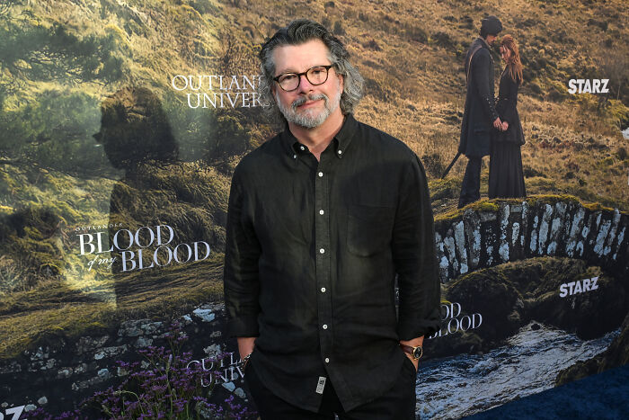 Man with glasses and gray hair stands in front of an Outlander Blood of My Blood promotional backdrop.