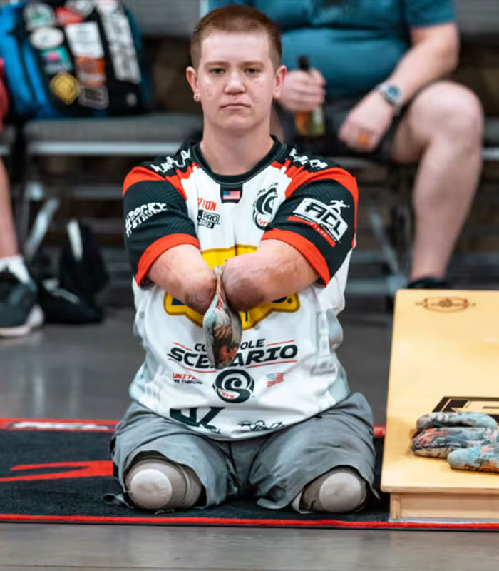 Man with no arms or legs wearing a sports jersey sitting on the floor, preparing to throw bags in a cornhole game.