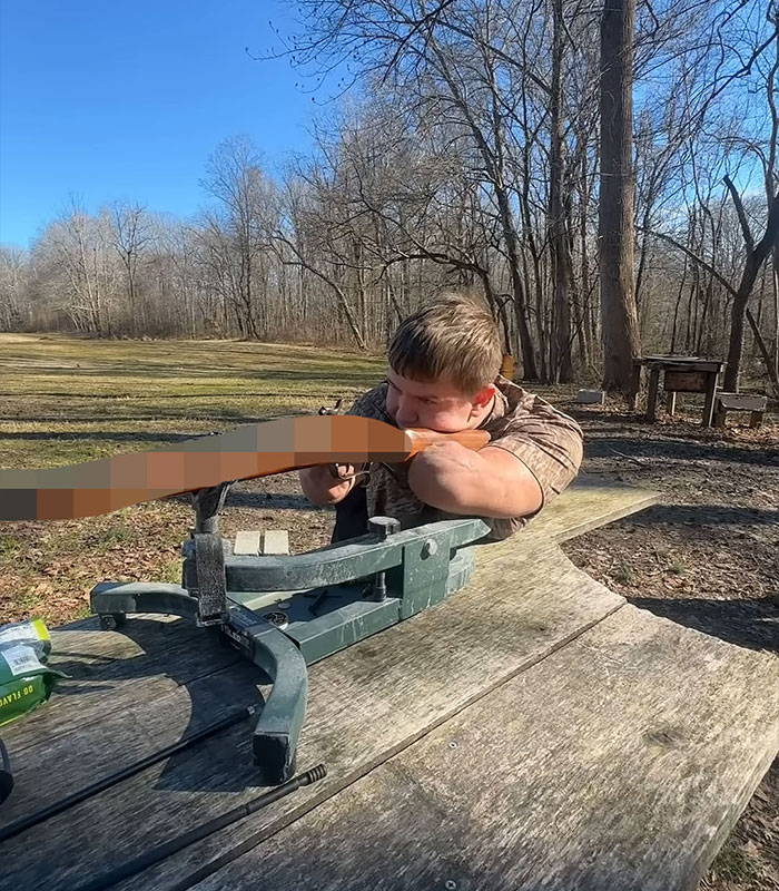 Young man aiming a rifle from a bench rest outdoors on a sunny day with bare trees in the background.