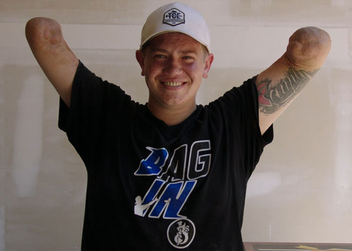 Man with no arms or legs smiling, wearing a white cap and black shirt with tattoo on his upper arm.