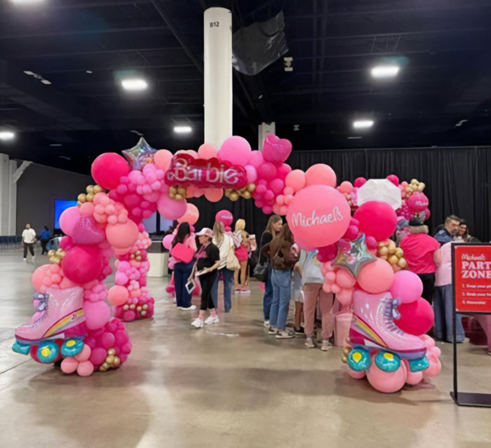Colorful pink balloon arch with Barbie and Michaels logos at Barbie Dream Fest event with people gathered inside venue.