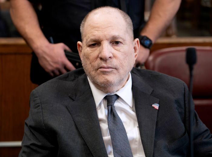Harvey Weinstein in a dark suit and tie, seated in a courtroom setting with security personnel in the background.