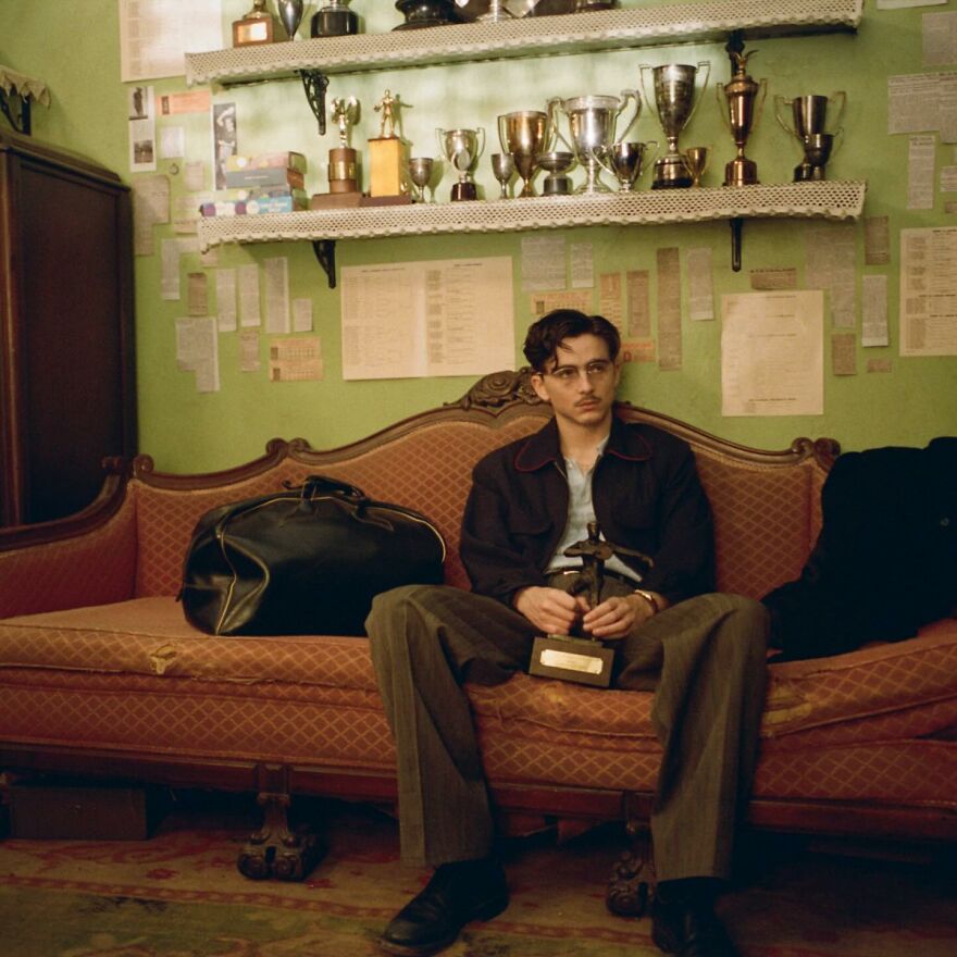 Young man holding award trophy on a vintage couch with Oscar-winning visuals and trophies displayed behind him