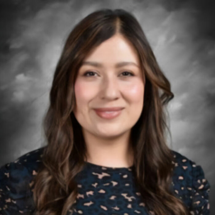 Portrait of a middle school secretary with long hair, smiling softly against a blurred gray background.