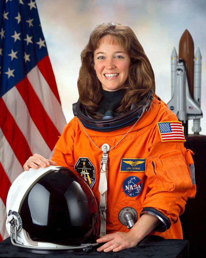 Female astronaut in an orange NASA suit with American flag, smiling and posing with a space helmet and shuttle model behind her.
