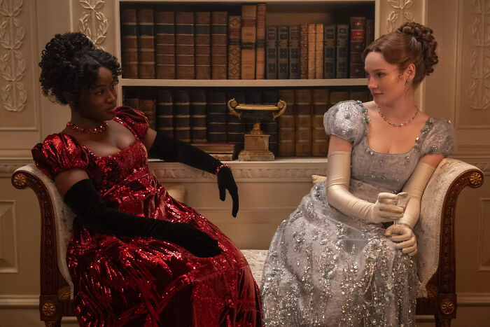 Two women in elegant historical dresses sit on a couch in a classic library setting discussing Bridgerton Season 5.