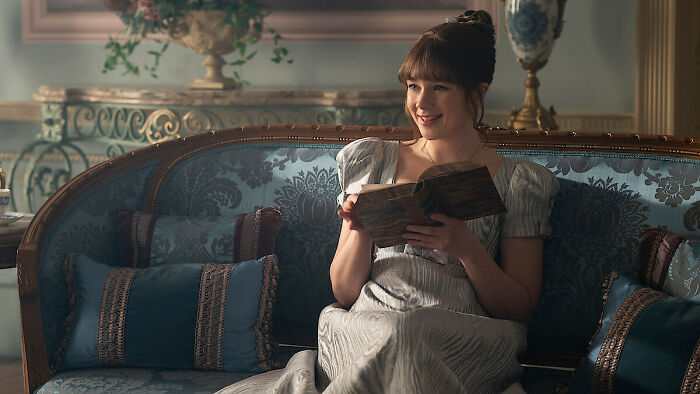 Young woman in period costume smiling while reading a book on an ornate couch, Bridgerton Season 5 update scene.