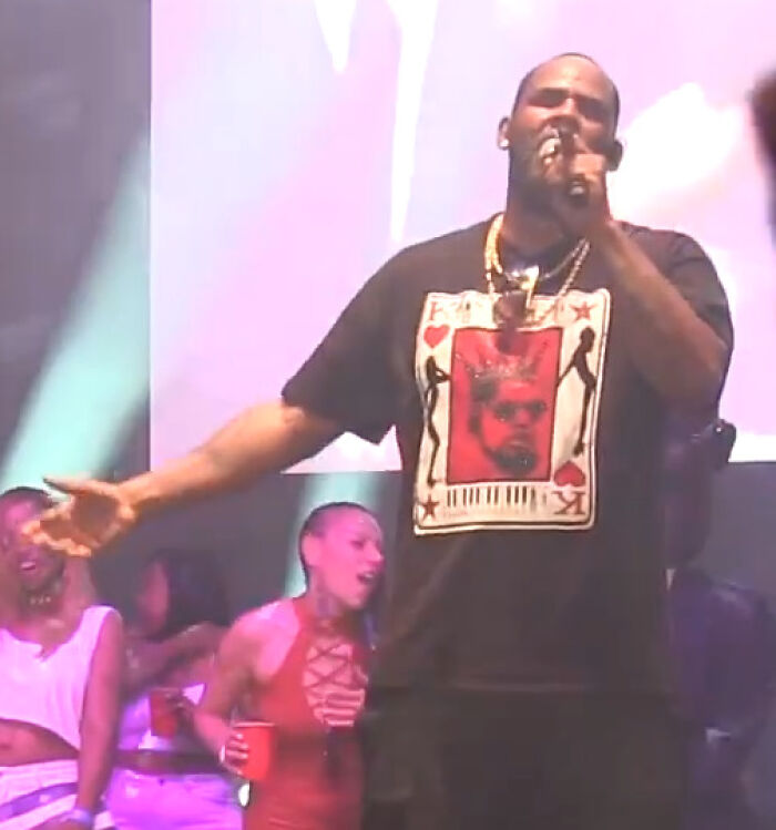 Man wearing a graphic t-shirt performing on stage with a microphone, with people dancing and holding cups nearby, celebrities hate.