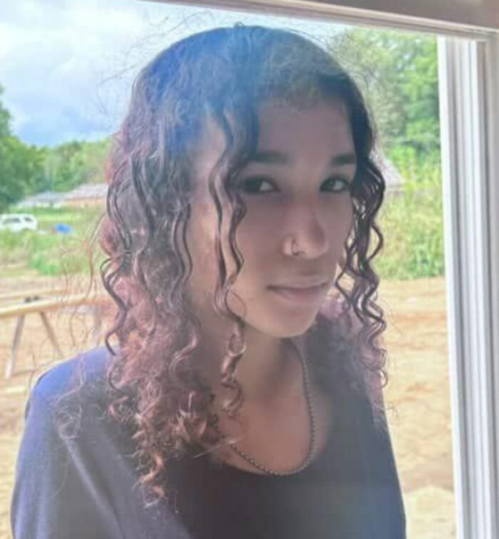 Young woman with curly hair standing near a window, related to bodies of father and daughter found on gravel road case.