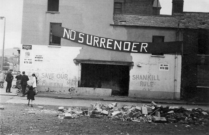 Black and white image of a street scene with a large no surrender banner, depicting a scary experience of conflict.