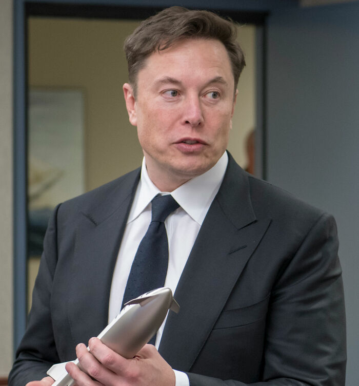Elon Musk holding a model rocket, dressed in a black suit and tie, highlighting odd details inside his house.