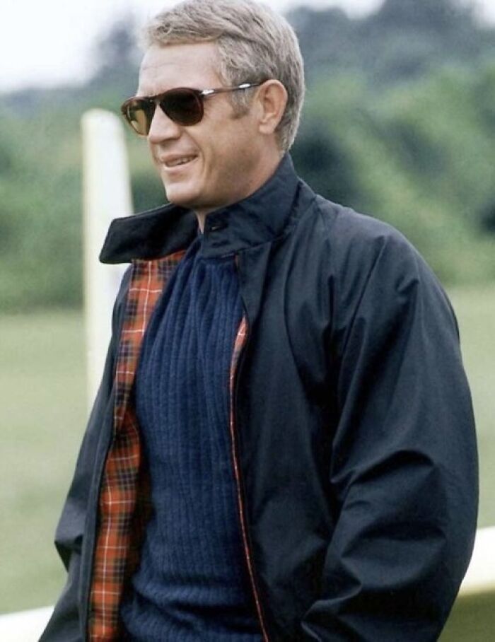 Man wearing sunglasses and a navy sweater under a jacket standing outdoors near a green field and a white post.