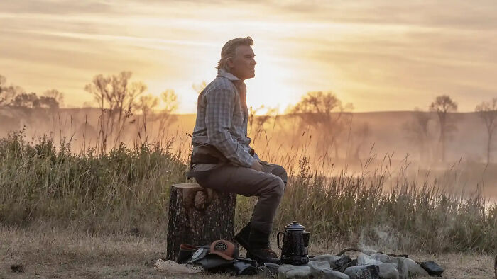 Man sitting on a log at a campfire in a rural setting, representing Dutton Ranch and Yellowstone successor series.