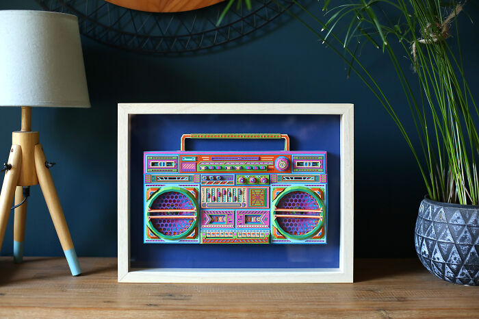 Colorful handcrafted paper boombox art by French artist duo displayed in a wooden frame on a home table.