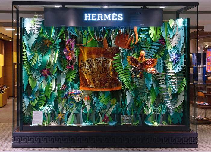Hermès store window display featuring handcrafted paper jungle scene with tiger and colorful birds by French artist duo.