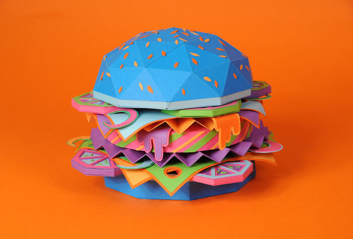 Colorful handcrafted paper burger created by French artist duo, showcasing intricate layers and vibrant design on an orange background.