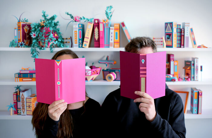 French artist duo holding pink handcrafted paper books with colorful paper art and models displayed on shelves behind them.