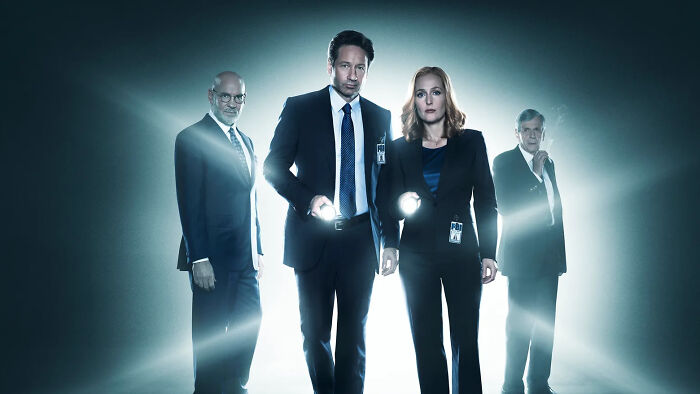Four cast members from The X-Files reboot, dressed in FBI attire, holding flashlights in a dark, misty setting.