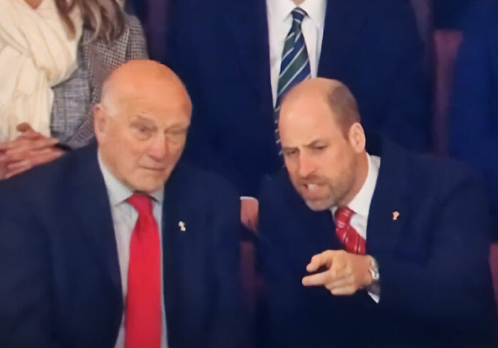 Prince William in a suit pointing and talking to an older man in a red tie, captured on camera going viral.
