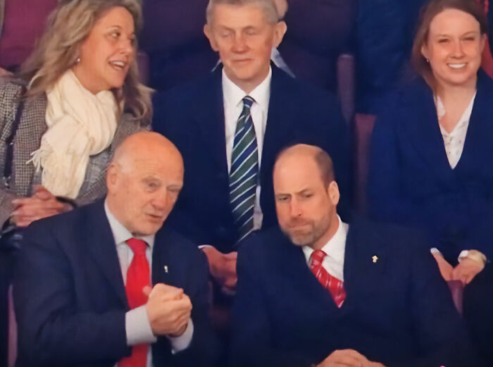 Prince William sitting in a crowd, wearing a suit and red tie, looking focused while engaging in conversation.