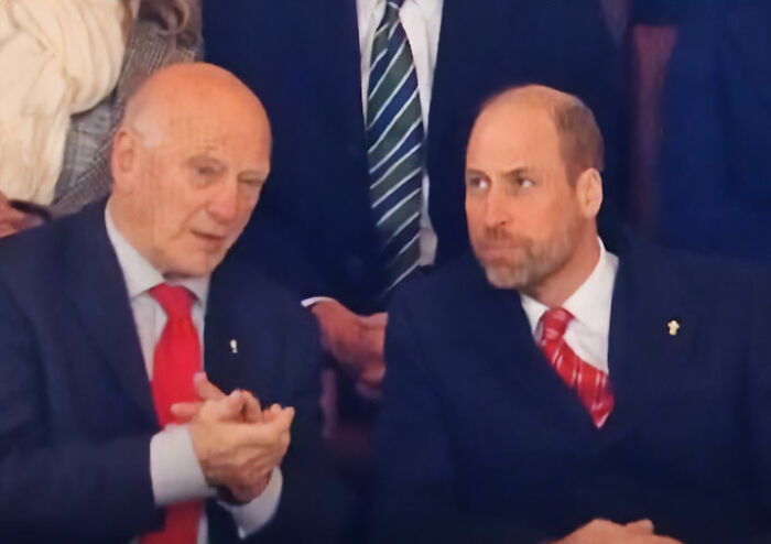 Prince William in a dark suit and red tie caught on camera getting into character during a formal event conversation.