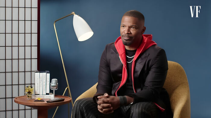 Man in a suit with red hoodie sitting in a chair, with a lamp and books on a side table in a studio setting
