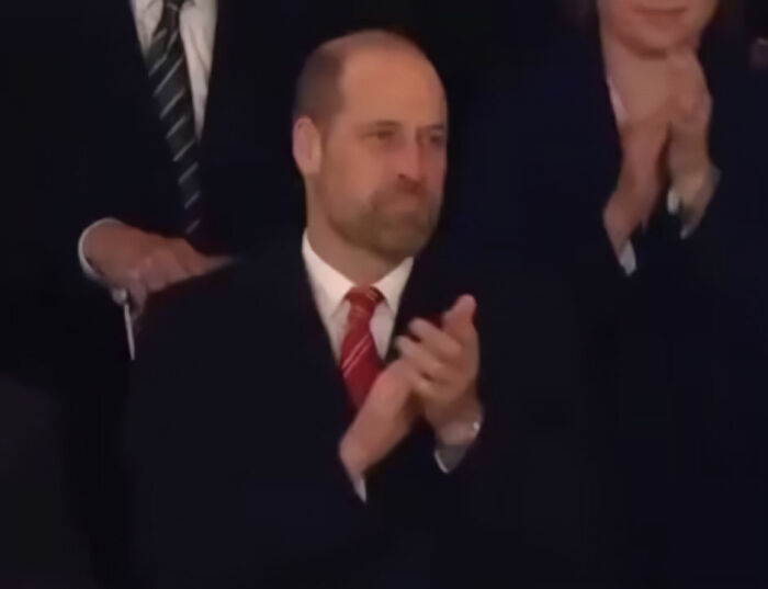 Prince William clapping in a dark suit and red tie, caught on camera during a viral moment getting into character.