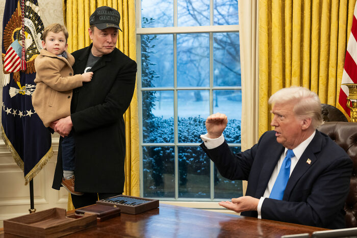 Elon Musk holding a child in a black coat and hat inside a room with yellow curtains, opposite Donald Trump at a desk.