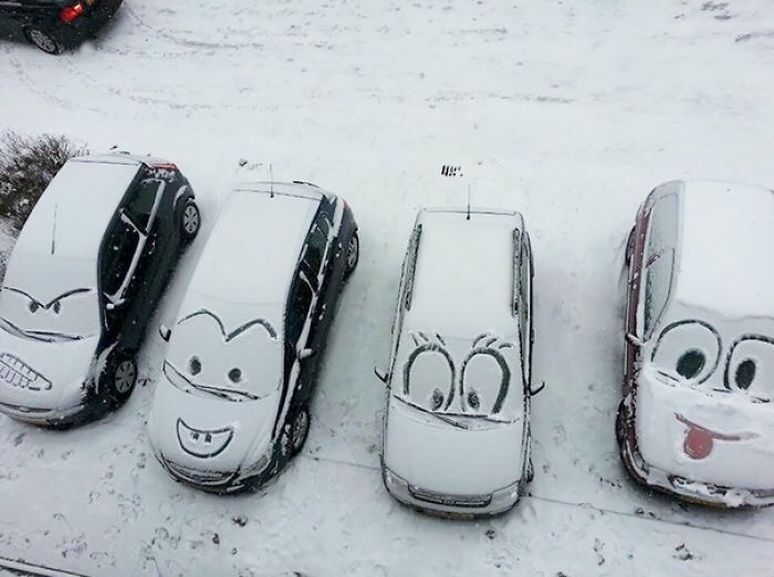 Four cars covered in snow with vandalism art depicting funny faces drawn on the windshields in a snowy parking lot.