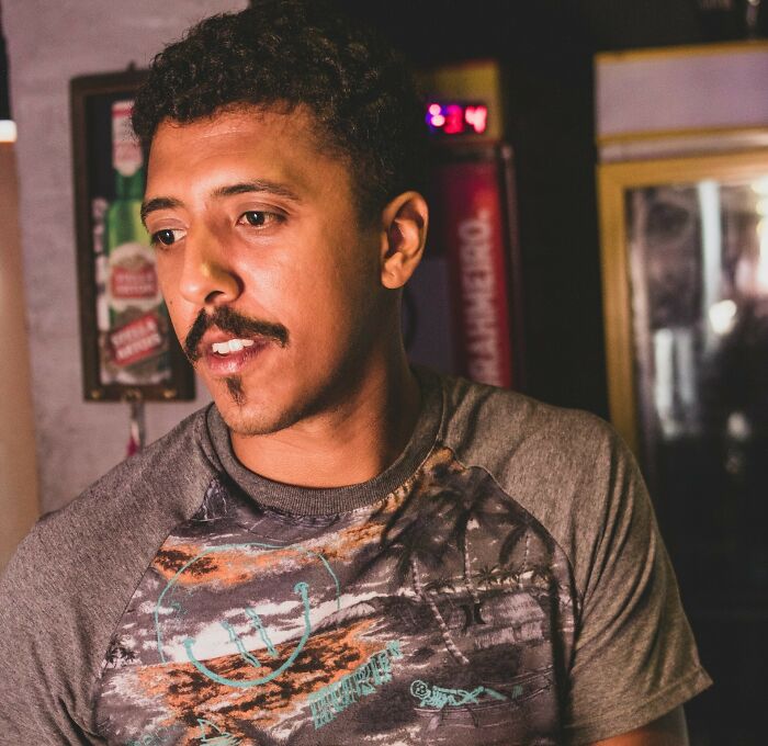 Young man with mustache and goatee, wearing a patterned shirt, in a dimly lit indoor setting, representing creepy advances context.