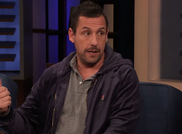 Actor Adam Sandler wearing a gray shirt and dark jacket during a TV interview discussing Gene Simmons and internet reactions.