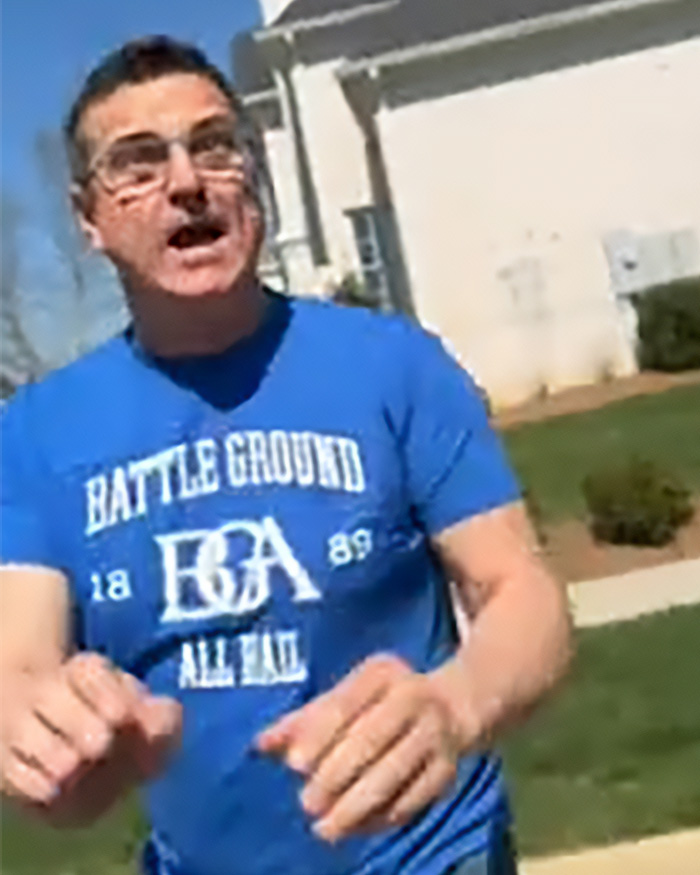 Man wearing blue Battle Ground t-shirt gesturing outdoors near a house, relating to Alan Ritchson wild fight with neighbor.