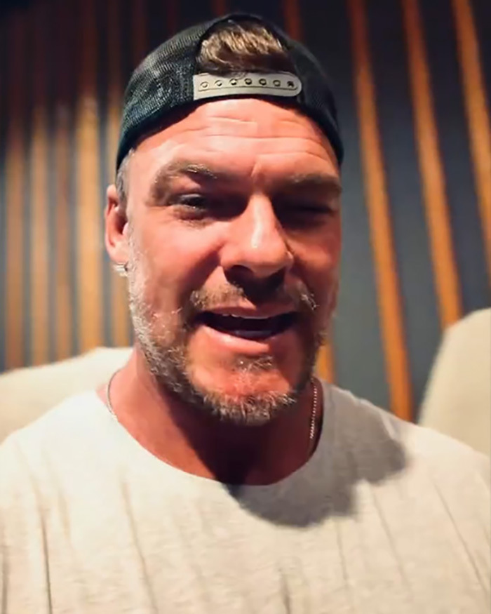 Alan Ritchson wearing a backward cap and white shirt, speaking about wild fight with neighbor in an indoor setting.