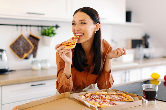 Young woman eating pizza in a bright kitchen, illustrating picky eater secretly enjoying regular food at home.