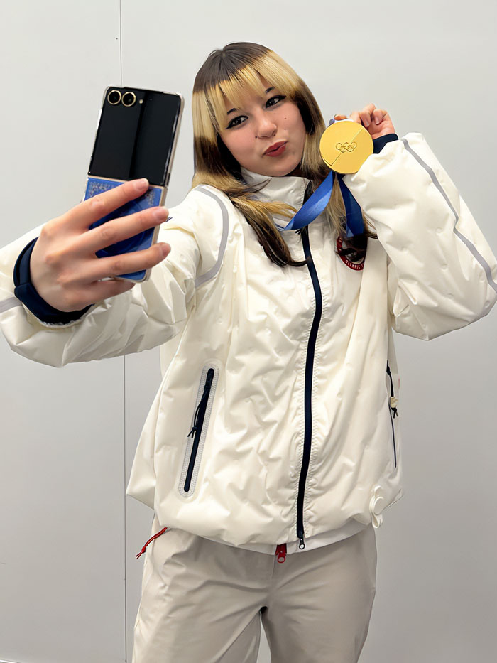 Olympic hero Alysa Liu wearing white jacket, holding gold medal and taking a selfie with a smartphone.