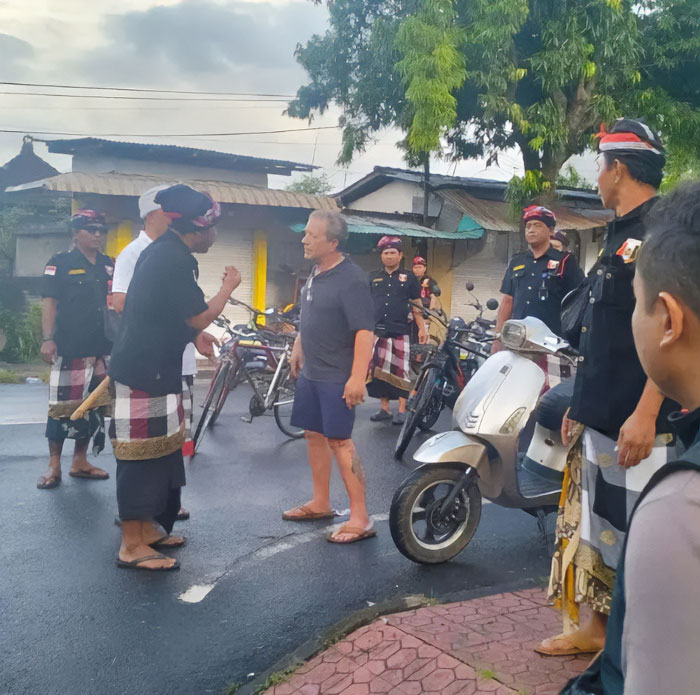 American tourist confronted by local authorities in Bali during public shutdown enforcement, surrounded by motorbikes and traditional attire.